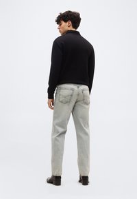 Νέος άνδρας γυρισμένος με την πλάτη, φοράει μαύρο μπλουζάκι με μακριά μανίκια, ανοιχτόχρωμα γκρι τζιν με πίσω τσέπες και μαύρες μπότες με τακούνι σε απλό φόντο.