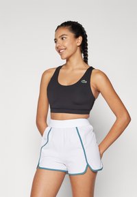 Svart sport-bh med rundad halsringning och grön logotyp; i kombination med vita shorts med turkos kantband och en elastisk midjeband.