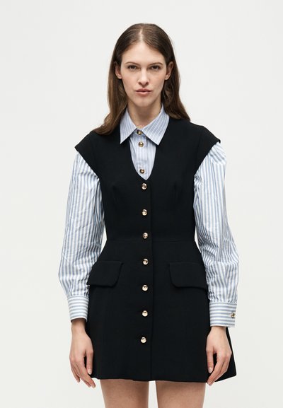 Femme portant une robe noire sans manches à boutons par-dessus une chemise à manches longues rayée bleu et blanc, debout devant un fond blanc.