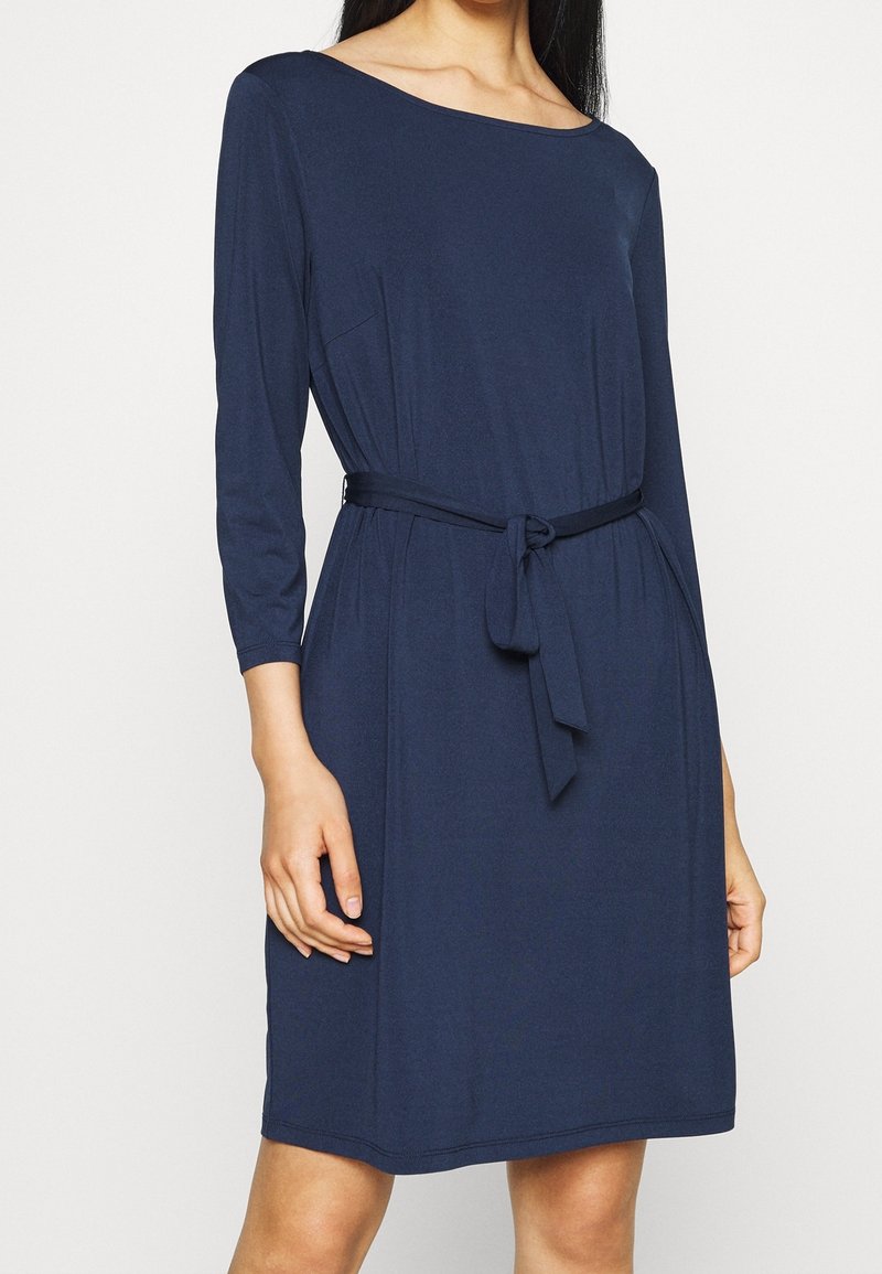 Femme portant une robe bleu marine longueur genou avec des manches trois-quarts et une ceinture nouée à la taille, debout devant un fond clair uni.