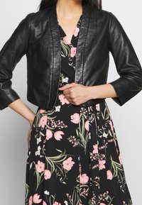 Černá kožená zkrácená bunda s hladkou texturou, vybavená mandarínským límcem a jemným prošíváním, vrstvená přes květinovou šaty.