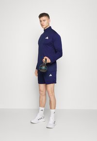 Tricou sportiv de culoare bleumarin cu guler înalt, mâneci lungi și logo alb, asortat cu pantaloni scurți. Modelul ține în mână o kettlebell neagră.