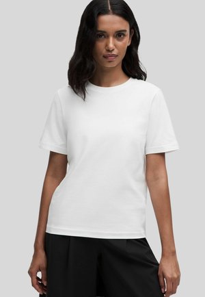 Frau mit langen dunklen Haaren, die ein schlichtes weißes T-Shirt mit kurzen Ärmeln und schwarze Hose trägt, steht vor einem hellgrauen Hintergrund.
