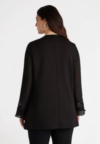 Tunik nera a maniche lunghe con dettagli in pizzo sulle maniche, tessuto morbido, taglio dritto e design minimalista. Si sente morbida e leggera.