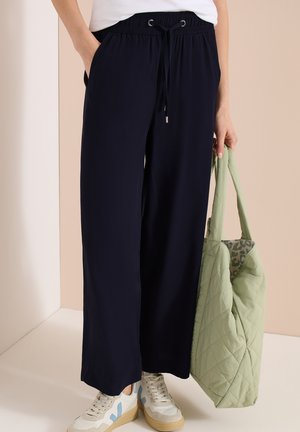 Osoba nosząca luźne czarne spodnie ze sznurkiem i białe trampki z beżowymi podeszwami, trzyma jasnzie zieloną pikowaną torbę na ramię.