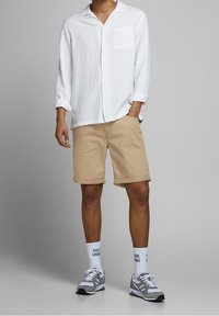 Camisa blanca de botones con mangas largas, pantalones cortos beige, zapatillas grises y calcetines blancos con texto. Ajuste casual y relajado; tela ligera.