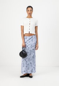 Haut blanc côtelé à manches courtes avec détail de boutons, associé à une jupe maxi en tie-dye bleu. Modèle tenant un petit sac à main noir, portant des chaussures plates.