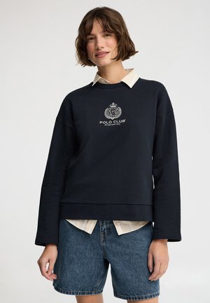 Mujer con cabello corto y castaño que lleva una sudadera azul marino Polo Club sobre una camisa beige con cuello y pantalones cortos de mezclilla, de pie frente a un fondo liso.
