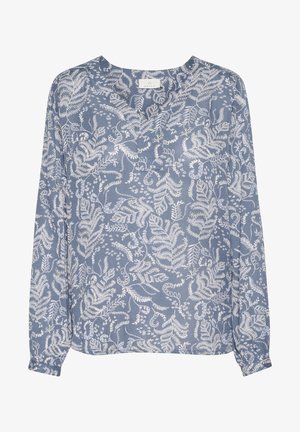 Gros plan sur une personne portant un haut bleu clair avec des motifs floraux et de feuilles blancs complexes, présentant un col en V et des manches longues.