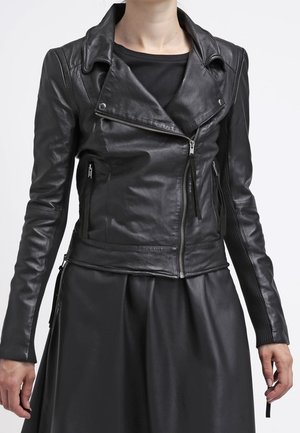 Veste en cuir noire asymétrique à fermeture éclair, portée sur un haut noir et associée à une jupe noire.