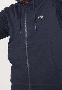 Sudadera con cremallera en color marino, confeccionada en tela suave, con un bolsillo tipo canguro, capucha ajustable y un pequeño logo de cocodrilo verde en el pecho.