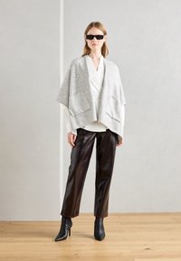 Grå uldponcho med to frontlommer over en hvid bluse, parret med skinnende brune bredde bukser og sorte spidse ankelstøvler.