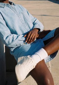 Personne portant une chemise en denim bleu clair et un short, assise les jambes croisées, portant des baskets blanches et des chaussettes sur une surface en béton.