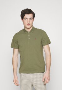 Olivengrønn polo skjorte laget av bomull, med krage, tre knapper, og et lite brodert emblem på brystet. Bært sammen med rutete bukser.
