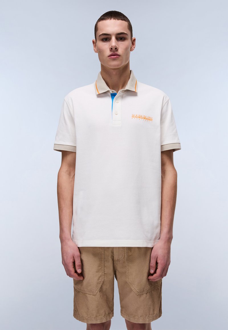 Polo shirt crema con accenti arancioni e blu, con tre bottoni e logo ricamato. Indossato con pantaloni corti beige, tessuto testurizzato.