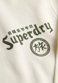 Sudadera blanca con texto verde "Superdry" en una fuente negrita y estilizada, con un gráfico circular adicional y caracteres japoneses.