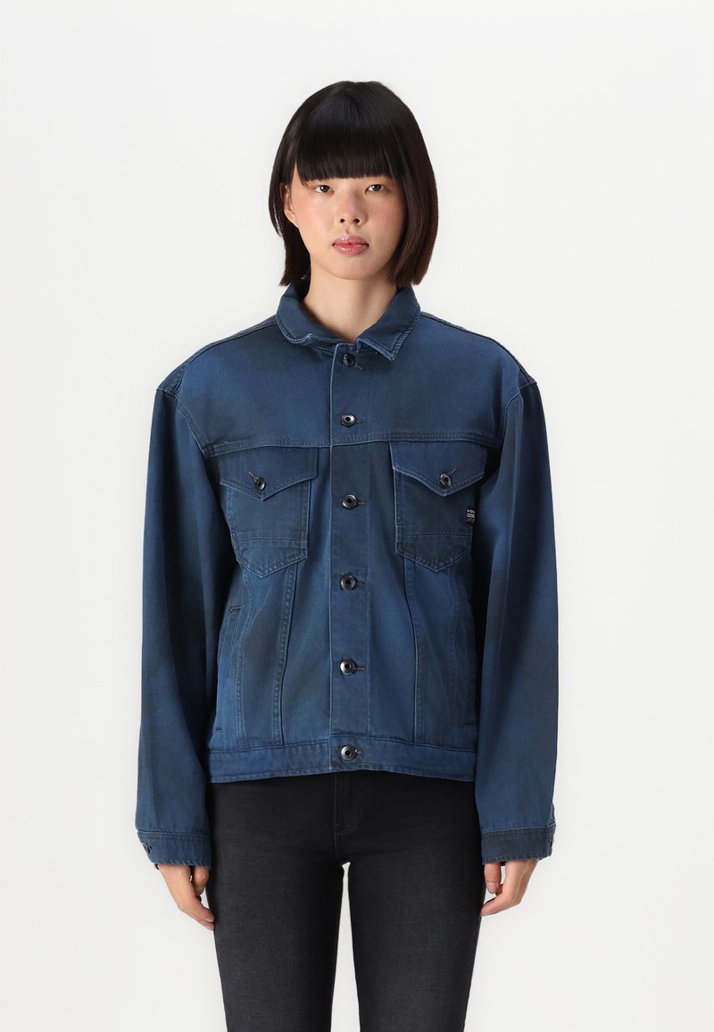 Τζάκετς από τζιν σε σκούρο μπλε χρώμα, με κουμπωτή μπροστινή όψη, δύο τσέπες στο στήθος και κλασικό γιακά. Έχει μακριά μανίκια και ελαφρώς υπερμεγέθη εφαρμογή.