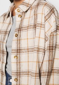 Donna che indossa una camicia a quadri beige e marrone con bottoni sopra un top grigio e jeans blu, vista dalla spalla alla vita.