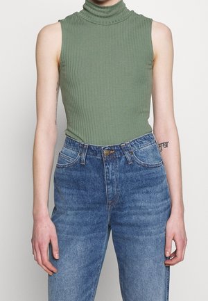 Zielony prążkowany bezrękawnik z golfem, wpuszczony w wysokowalne niebieskie jeansy, z widocznymi tatuażami na obu ramionach.