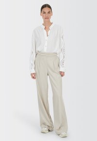 Weißes Hemd mit Knopfverschluss und Spitze an den Ärmeln, kombiniert mit hochgeschnittenen, beigen Nadelstreifenhosen und hellen Sneakern.