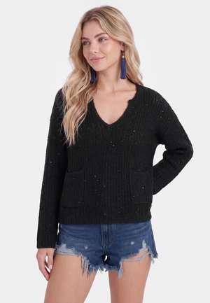Blonde vrouw met lang haar draagt een zwarte gebreide trui en blauwe versleten denim shorts, geaccentueerd met lange blauwe kwastjes oorbellen.