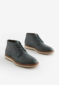 Botas chukka de cuero en gris oscuro con costuras azules, que presentan una puntera redonda, un cierre de cordones en la parte delantera y una suela beige contrastante.