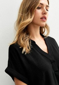 Top nero dalla vestibilità ampia con scollo a V, maniche corte arrotolate e un tessuto testurizzato, mostrato su uno sfondo neutro.