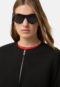 Czarne okulary przeciwsłoneczne z zaokrągloną oprawką i ciemnymi soczewkami, noszone z czarną kurtką z czerwonym kołnierzem i detalem na zamku błyskawicznym.