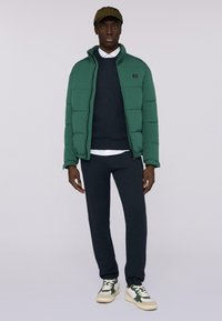 Zielona kurtka puchowa z wysokim kołnierzem, granatowy sweter, granatowe dresy oraz zielono-białe trampki. Model ma na sobie oliwkową czapkę.