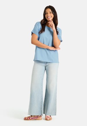Frau lächelnd, trägt ein hellblaues kurzärmeliges Oberteil mit Häkelärmeln, weite hellblaue Jeans und rosa gestreifte Sandalen, steht vor weißem Hintergrund.