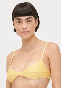 Keltainen bikinitoppi, jossa on kolmiomainen muoto, ohuet hihnat ja ristikkäinen muotoilu keskellä. Silkkinen pinta ja minimaalinen yksityiskohtaisuus korostavat yksinkertaisuutta.