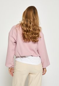 Svetloružová skrátená bunda s textúrovaným tkaním, širokými rukávmi, cez biely top, skombinovaná s béžovými nohavicami, zobrazená zozadu.