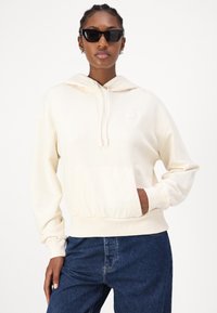 Cream-colored hoodie with a kangaroo pocket, hood with drawstrings, and ribbed cuffs; paired with blue denim jeans. Black rectangular sunglasses.