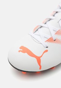 Scarpina da calcio bianca con superficie testurizzata, dettagli arancioni e un grande logo Puma. Presenta un design con lacci e tacchetti visibili sulla suola.