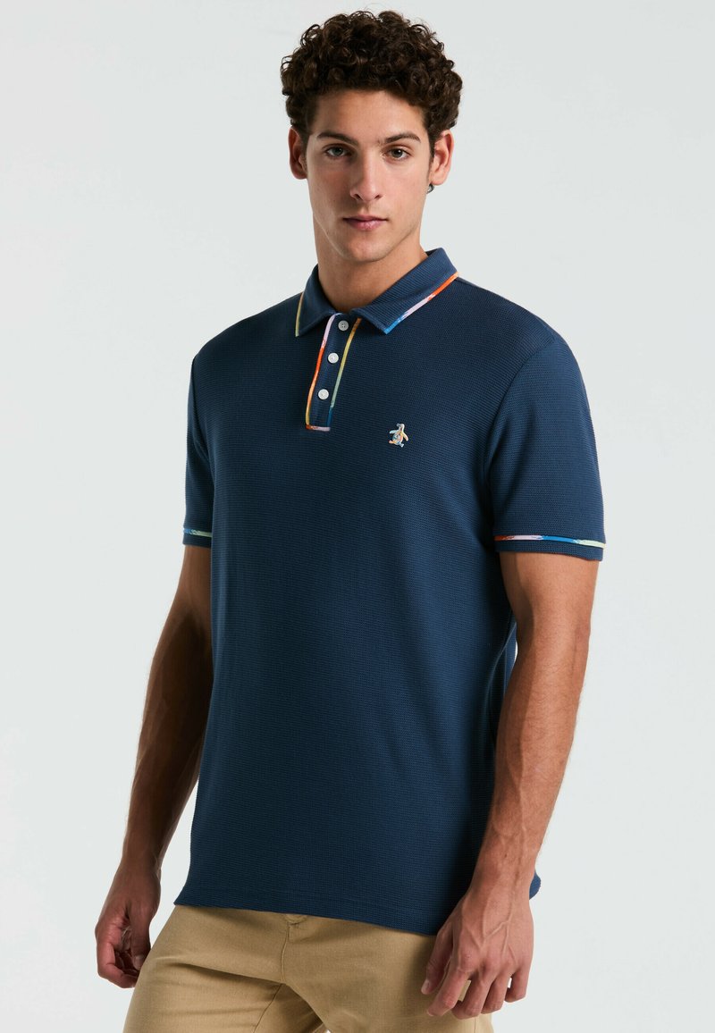 Polo bleu marine en tissu texturé, avec un col et des bords de manches colorés contrastants, une patte de boutonnage à trois boutons et un petit logo sur la poitrine.