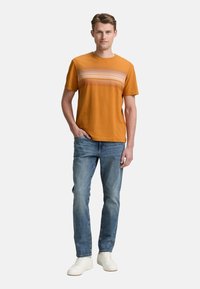 Kortærmet orange T-shirt med vandrette striber i lyse nuancer, parret med blå denimjeans og hvide sneakers.