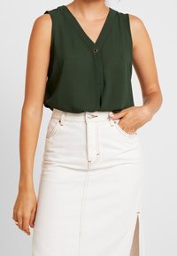 Blusa sin mangas de color verde oscuro con escote en V, un detalle de un solo botón, combinada con una falda vaquera blanca con una abertura lateral.