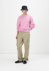 Sweatshirt rose avec un petit logo, pantalon cargo beige avec plusieurs poches, et baskets noires. Le mannequin porte un chapeau seau noir.