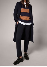 Manteau long navy sur un pull rayé marron et navy, chemise blanche à col, pantalon noir sur mesure et chaussures à plateforme noires.