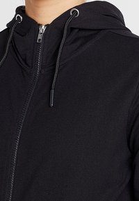 Sudadera con cremallera negra y capucha ajustable, con una tela suave y texturizada y costuras visibles a lo largo de la cremallera.