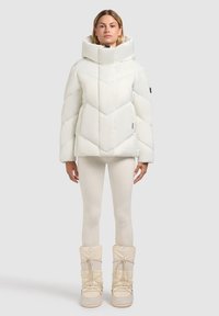 Hvid pufferjakke med høj krave og chevronmønster, parret med beige leggings og snesko med snørelukning.