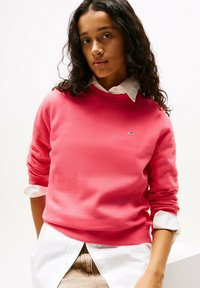 Felpa rosa con maniche lunghe e polsini a coste, indossata sopra una camicia bianca con colletto. Patch del logo ben visibile sul petto.