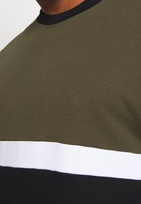 Camiseta verde oliva con una franja horizontal en blanco y negro. Cuenta con cuello redondo y est�á hecha de algodón suave y texturizado.