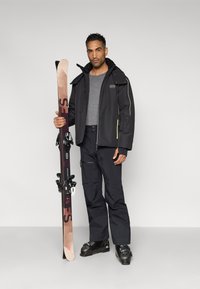 Μαύρο χειμερινό μπουφάν με κίτρινες λεπτομέρειες, γκρι πουκάμισο, μαύρο χειμερινό παντελόνι και μαύρα χειμερινά παπούτσια, κρατώντας σκι με δεσίματα. Απλός, λειτουργικός σχεδιασμός.