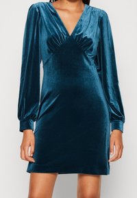 Tyrkysové sametové šaty s hlubokým výstřihem do V, nařasením v oblasti živůtku a dlouhými balónovými rukávy, se šitým siluetou a hladkým povrchem.