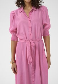 Robe rose à boutons avec manches bouffantes et poignets smockés. Elle présente une ceinture à la taille, une texture douce et une silhouette droite.