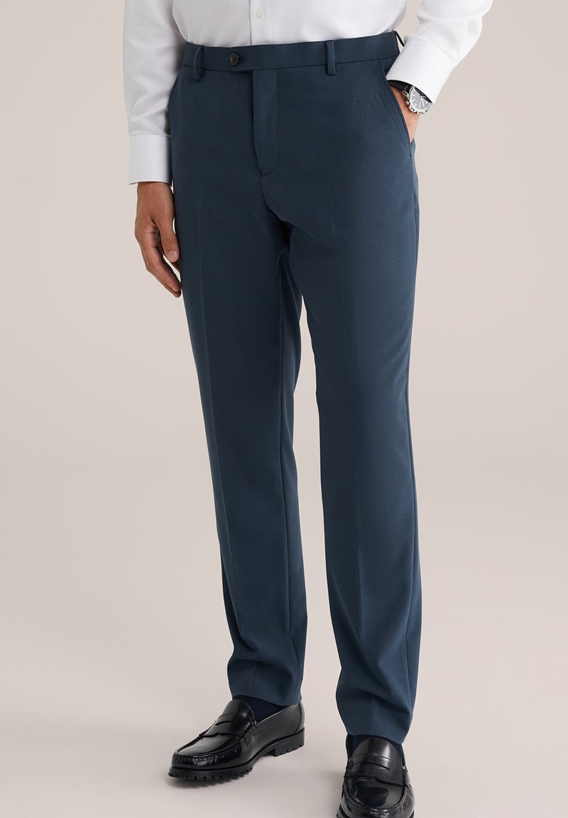 Pantalon de costume bleu marine avec une taille plate, deux poches latérales et une fermeture à un bouton, assorti à des mocassins noirs et une chemise blanche à boutons.