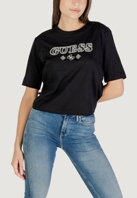T-shirt nera corta con logo "GUESS" in ricamo bianco e argento, abbinata a jeans aderenti azzurri. Materiale in cotone liscio.
