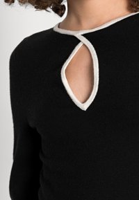 Svart stickad topp med vit kant runt en nyckelhålsöppning vid halsen, som har en slät textur och en figurnära design.