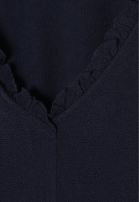 Tessuto blu navy con un motivo testurizzato, caratterizzato da una scollatura a V e un bordo arricciato lungo la scollatura per un tocco di dettaglio in più.